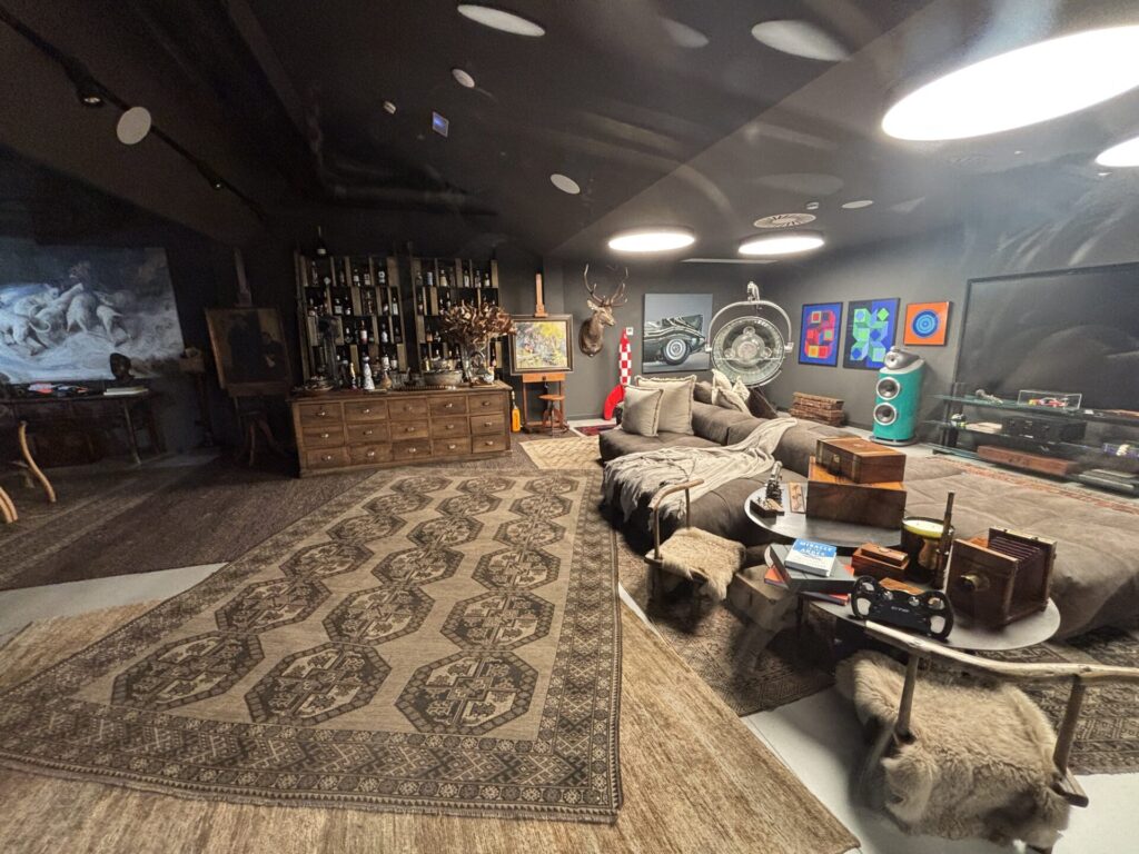 An eclectic meeting room with a huge rug, a huge sofa with a blanket and pillows with a lot of different random things in the background.